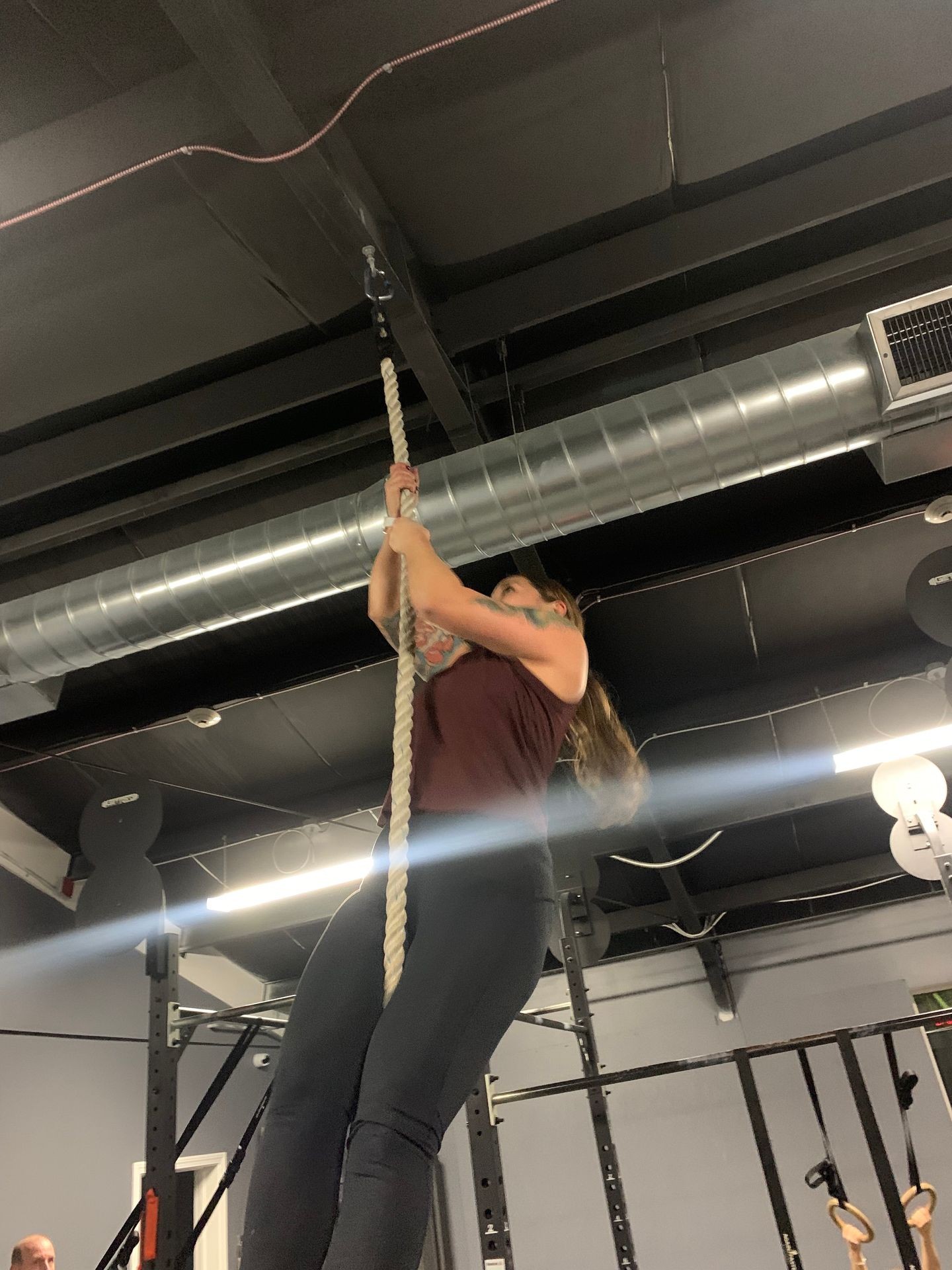 Person climbing a rope in a gym setting with industrial-style ceiling and workout equipment in the background.